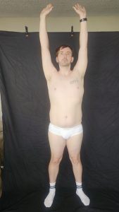 Joshua Marron facing forward with arms raised overhead in white briefs and socks during Project Legacy vegan body transformation.