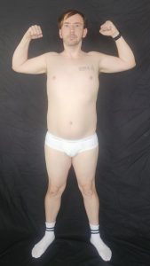 Joshua Marron flexing biceps facing forward in white briefs and socks, showing muscle progress for Project Legacy transformation.