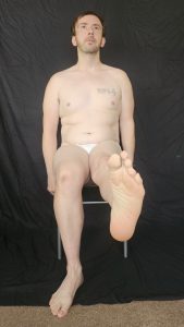 Joshua Marron seated with left leg extended forward flexing toes in white briefs for Project Legacy vegan fitness progress photo.